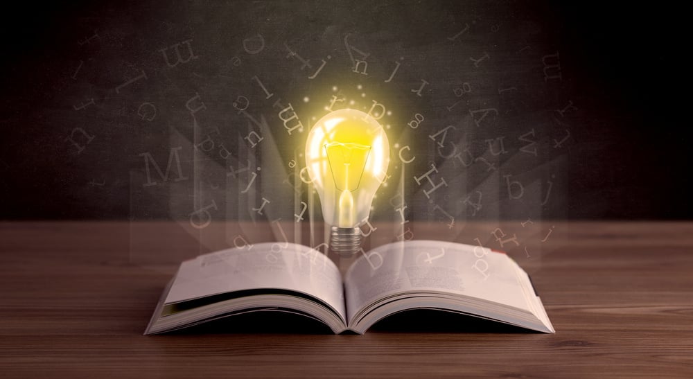 Shiny alphabet letters and yellow lightbulb hovering over open book